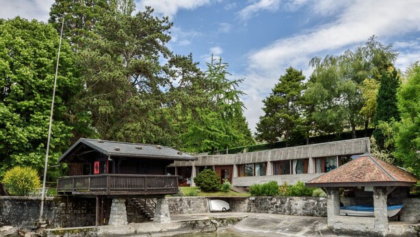 A rustic lodge with a wooden cabin and a concrete building, surrounded by lush greenery and a clear pool.