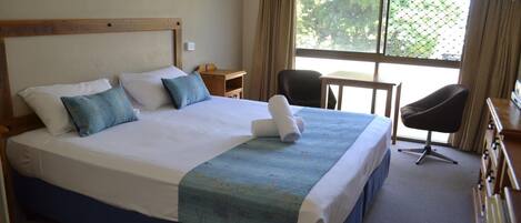 A large bed with a wooden headboard, a blue bedspread, and two pillows. There is a chair and a small table next to the bed.
