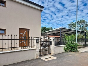 Um edifício com fachada bege, porta de madeira, portão de metal preto e área externa coberta.