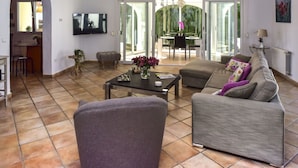Un salón amplio con sofá, sillón y mesa de centro. Se ve un televisor montado en la pared, y a través de las puertas de cristal se distingue una zona de comedor con mesa y sillas.