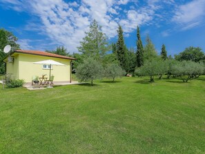 Uma casa amarela com telhado de cerâmica, um pátio com mesa e cadeiras e um gramado com árvores.