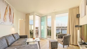 A modern living room with a grey sofa, a small table with a bowl of fruit, a grey armchair, and a TV on a stand. Large windows offer a view of a marina with boats.