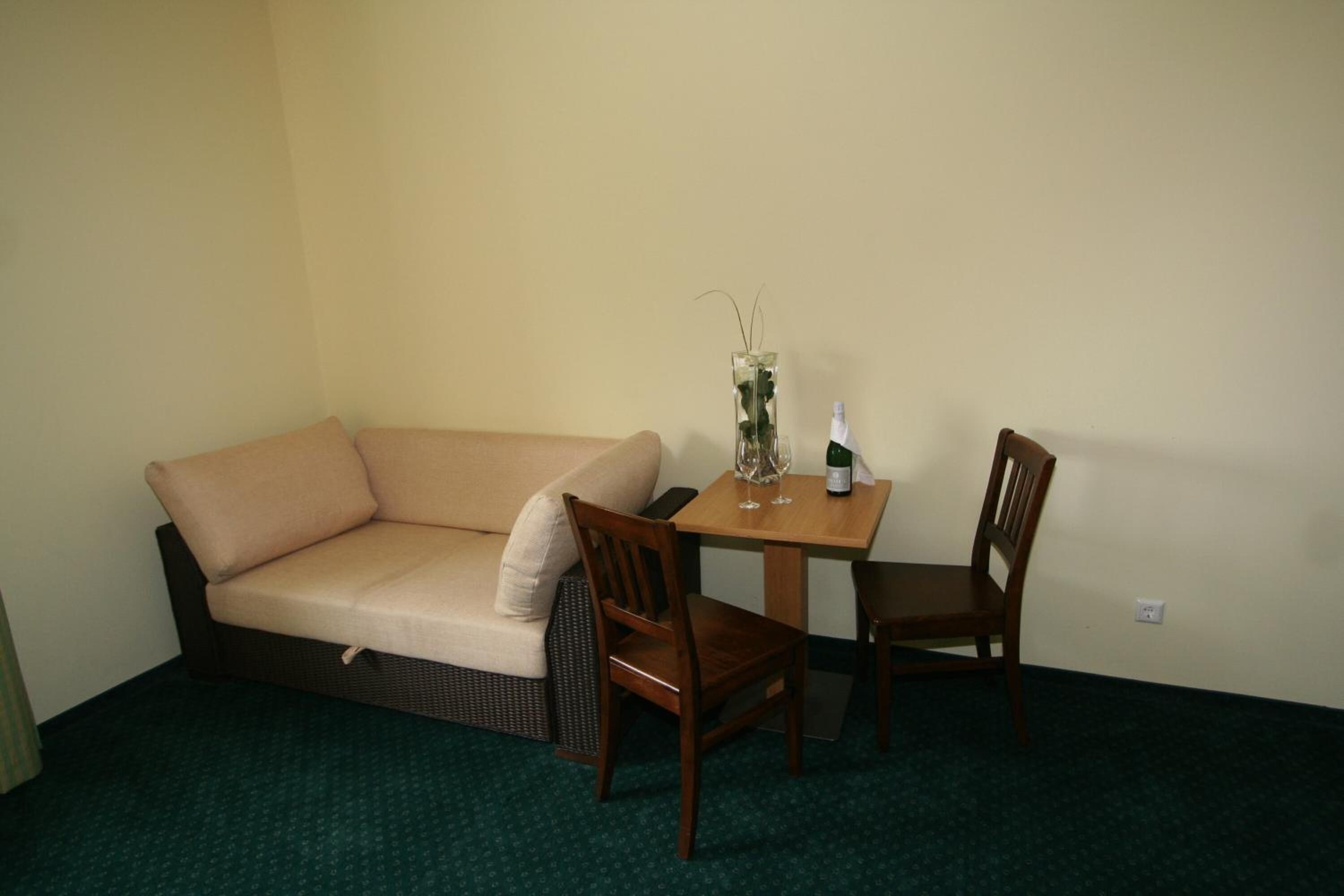 A small room with a beige sofa, a wooden table with two chairs, a vase with a plant, and a bottle.