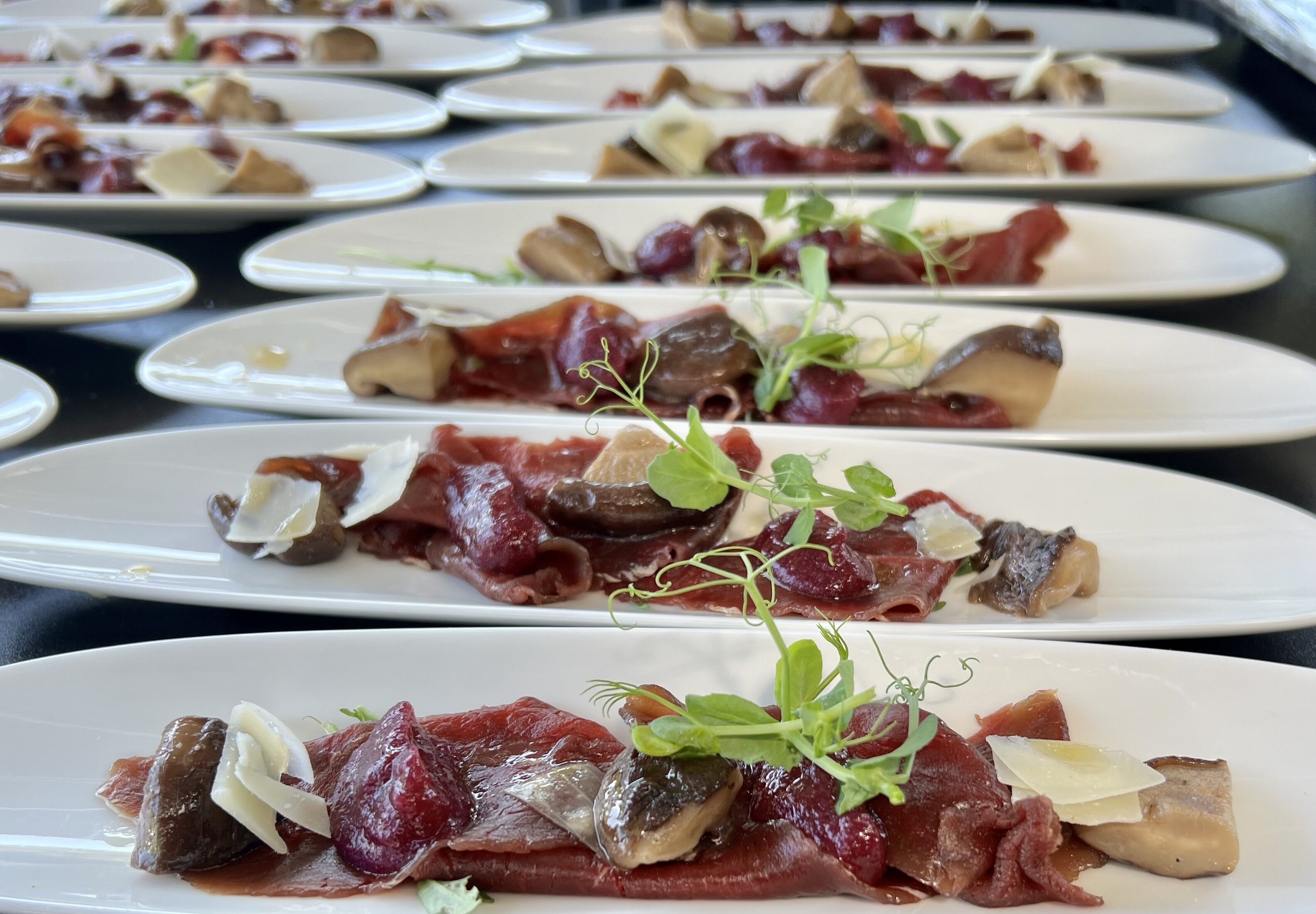 A row of white plates with food, each topped with mushrooms, meat, and greens.