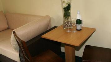 Una habitación pequeña con un sofá color beige, una mesa de madera con una botella y vasos, y un jarrón con flores.