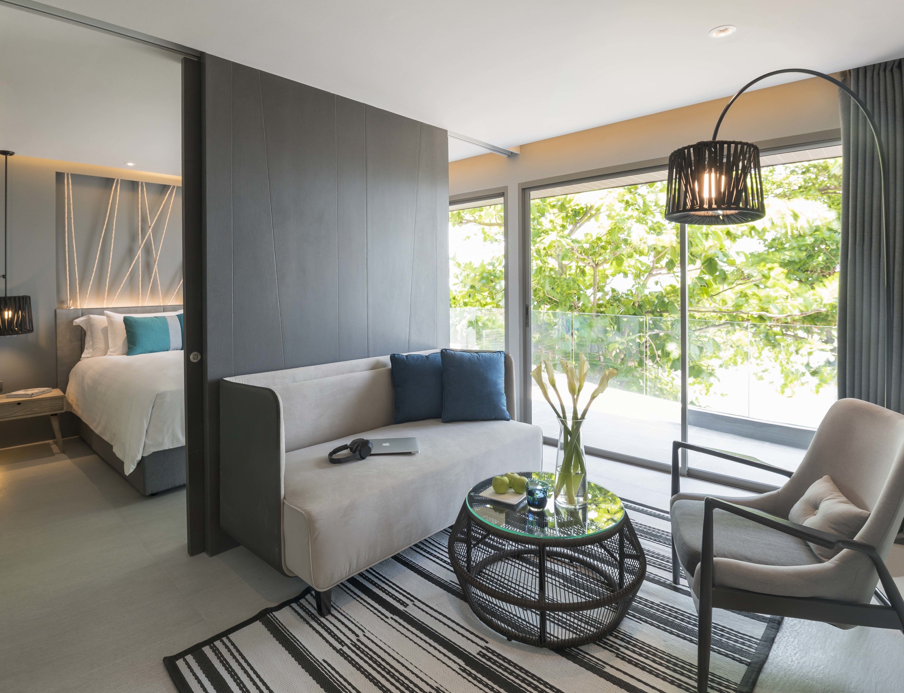 A modern hotel room with a sofa, a round coffee table, a chair, and a large window offering a view of greenery.