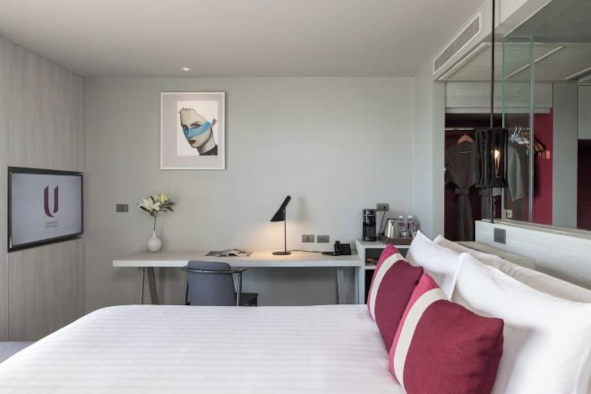 Moderni hotellihuone, jossa on siististi pedattu sänky, työpöytä lampun kanssa, pieni pöytä maljakossa olevilla kukilla sekä suuri peili, joka heijastaa huonetta.