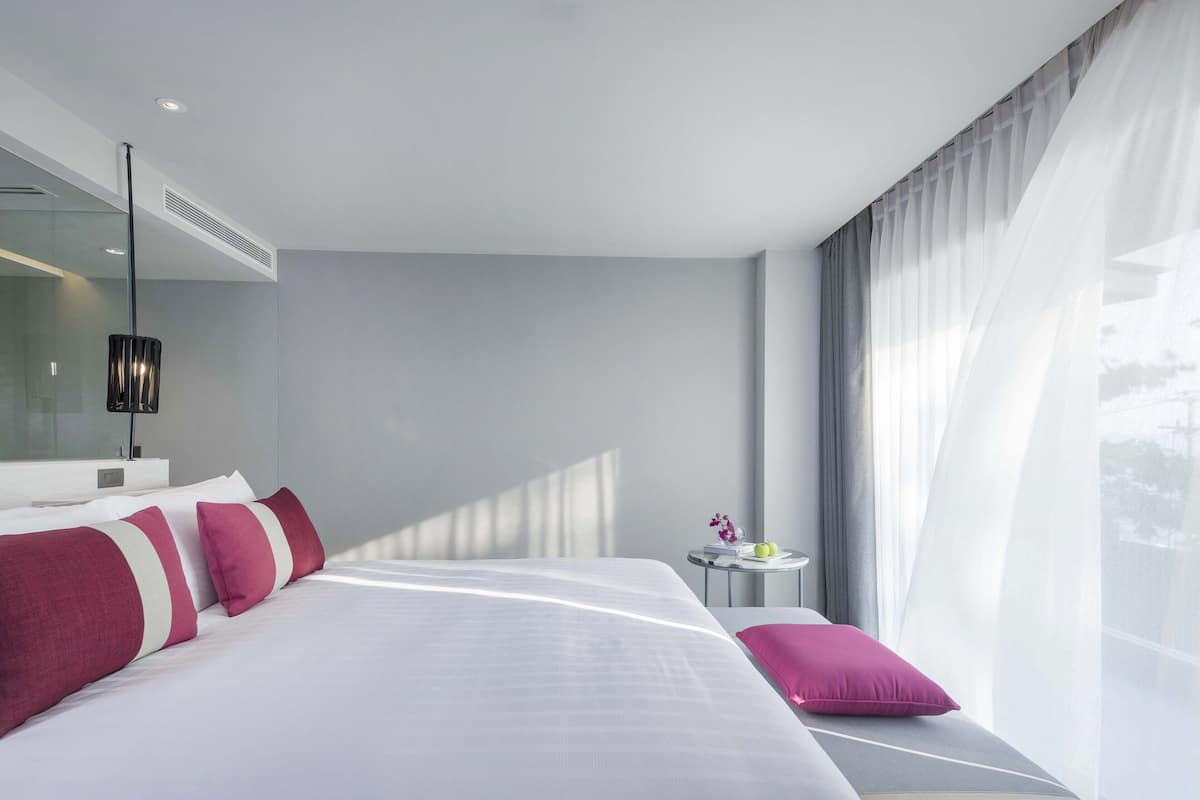 Una habitación de hotel moderna con una cama grande, una mesita de noche con lámpara y vistas al exterior a través de cortinas translúcidas.