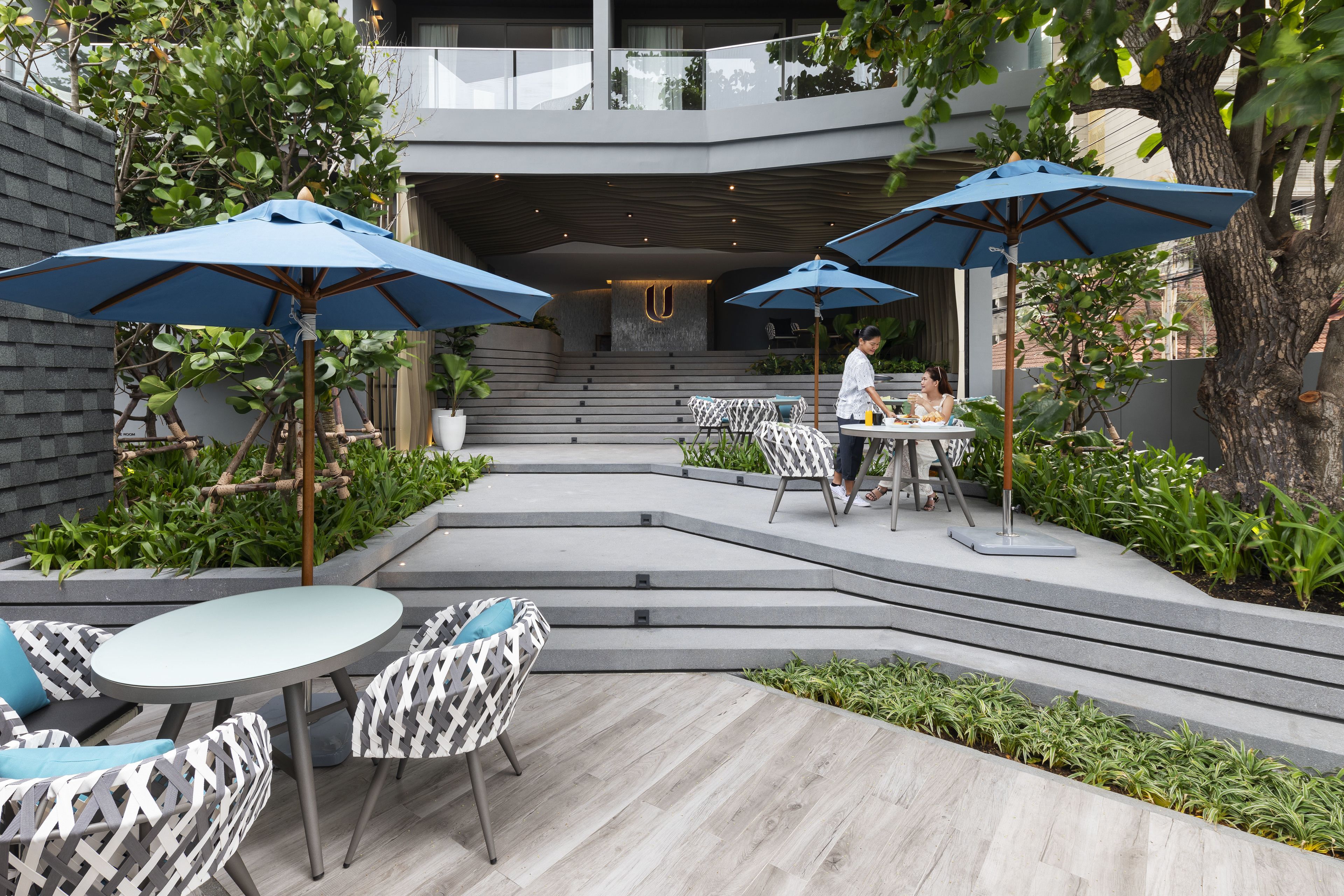An outdoor patio with modern furniture, blue umbrellas, and a staircase leading to a balcony.