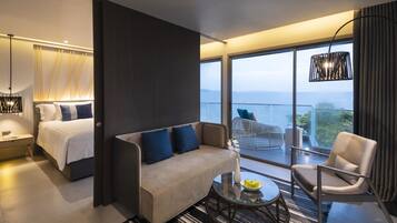 A modern hotel room with a bed, sofa, armchair, and a balcony with a view.