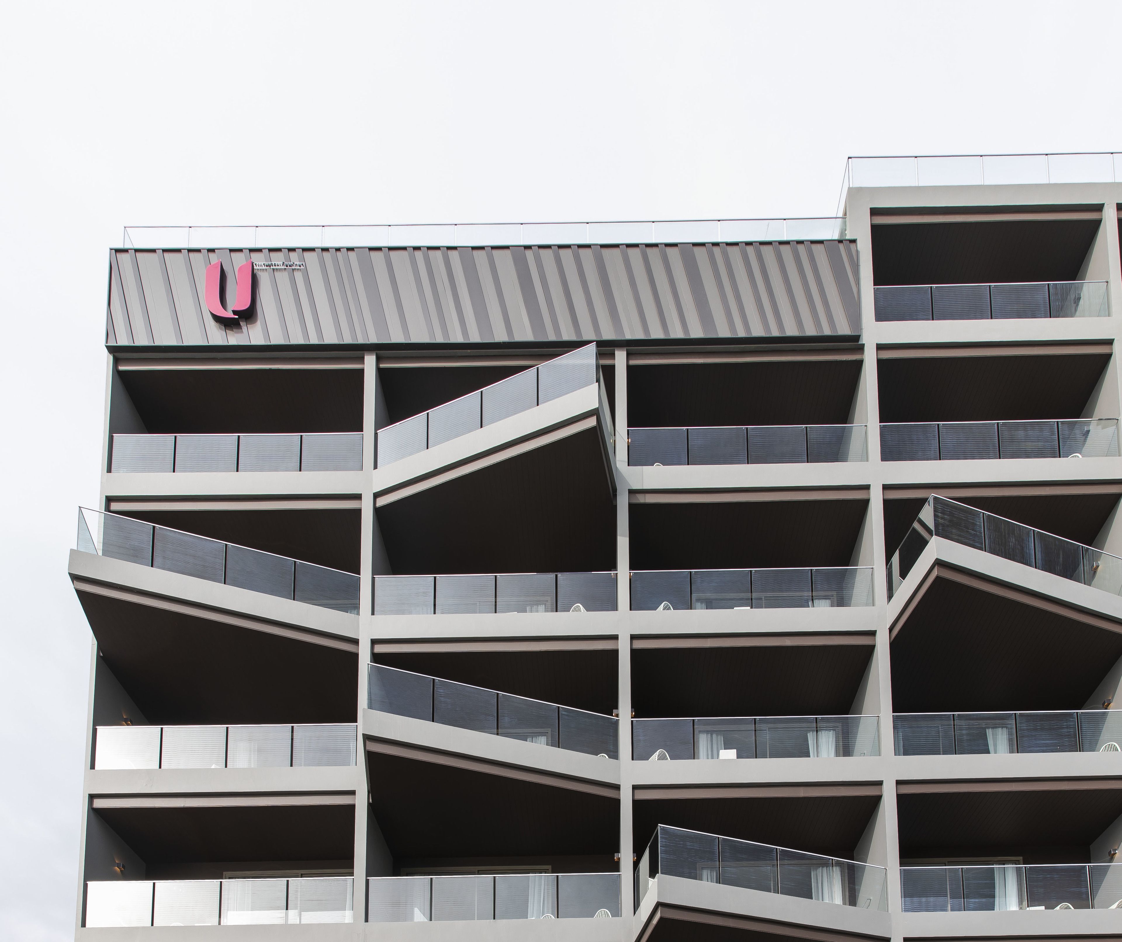 Een meerlaags gebouw met een modern architectonisch ontwerp, voorzien van balkons met glazen hekwerk en een opvallend logo bovenaan.