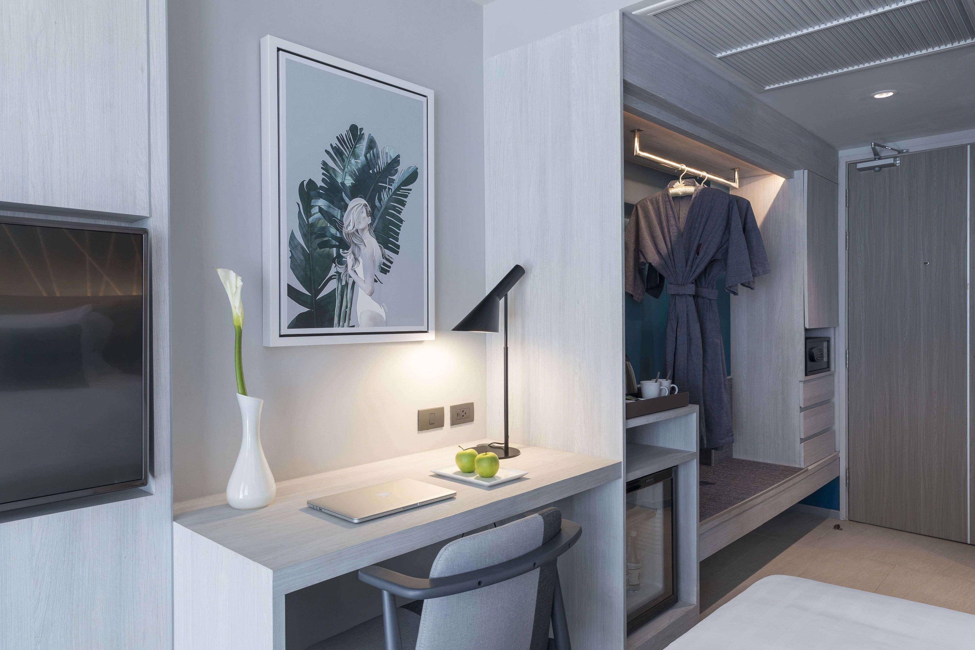 A modern hotel room with a desk, a flat-screen TV, a framed artwork, a vase with flowers, and a walk-in closet with hanging robes.
