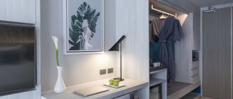 A modern hotel room with a desk, a flat-screen TV, a framed artwork, a vase with flowers, and a walk-in closet with hanging robes.