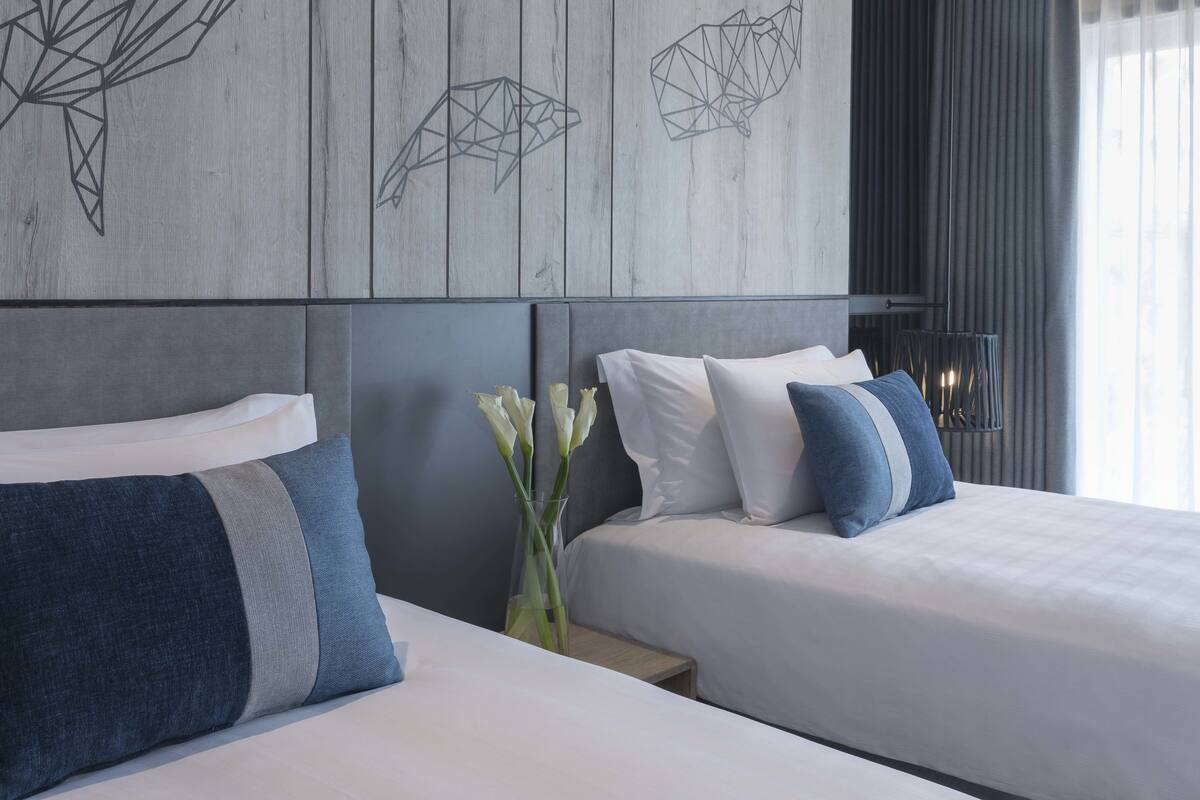 Habitación de hotel con dos camas, una mesita de noche, un jarrón con flores y decoración mural geométrica.