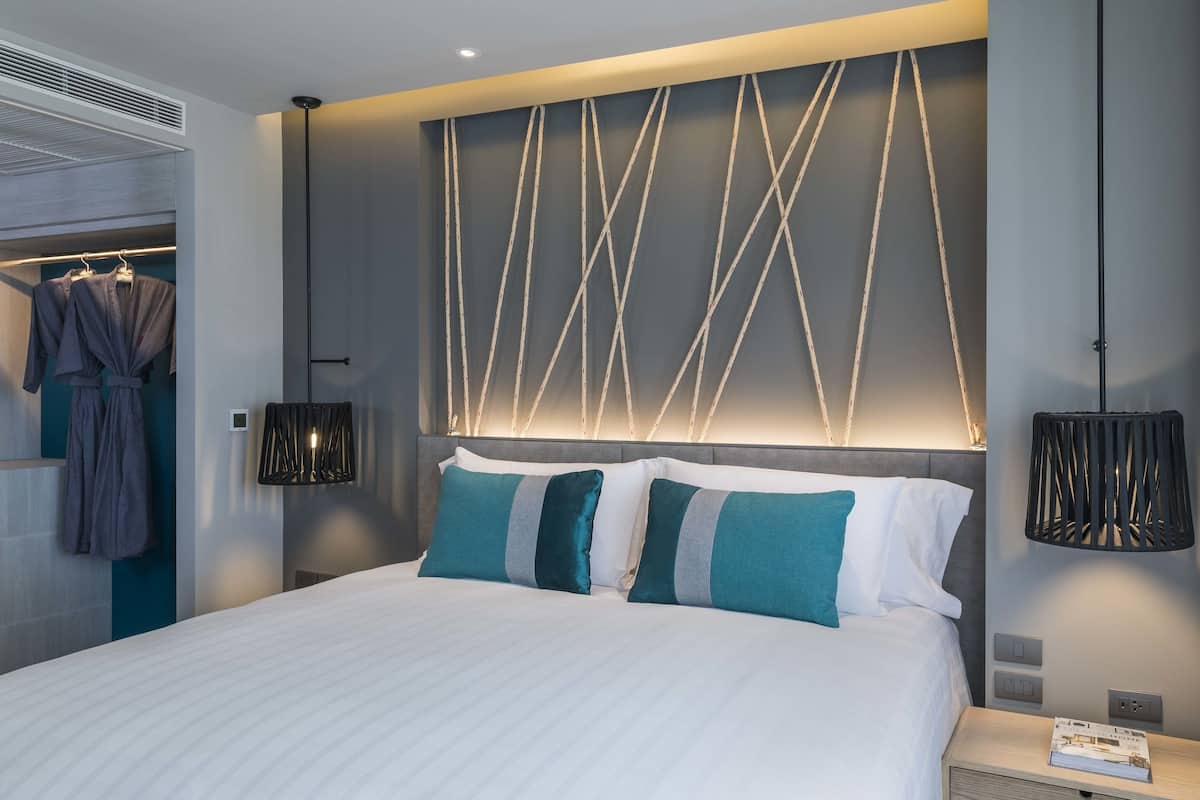 Una habitación de hotel moderna con una cama grande, un cabecero con un diseño de luz original, lámparas de noche y un armario con ropa colgada.