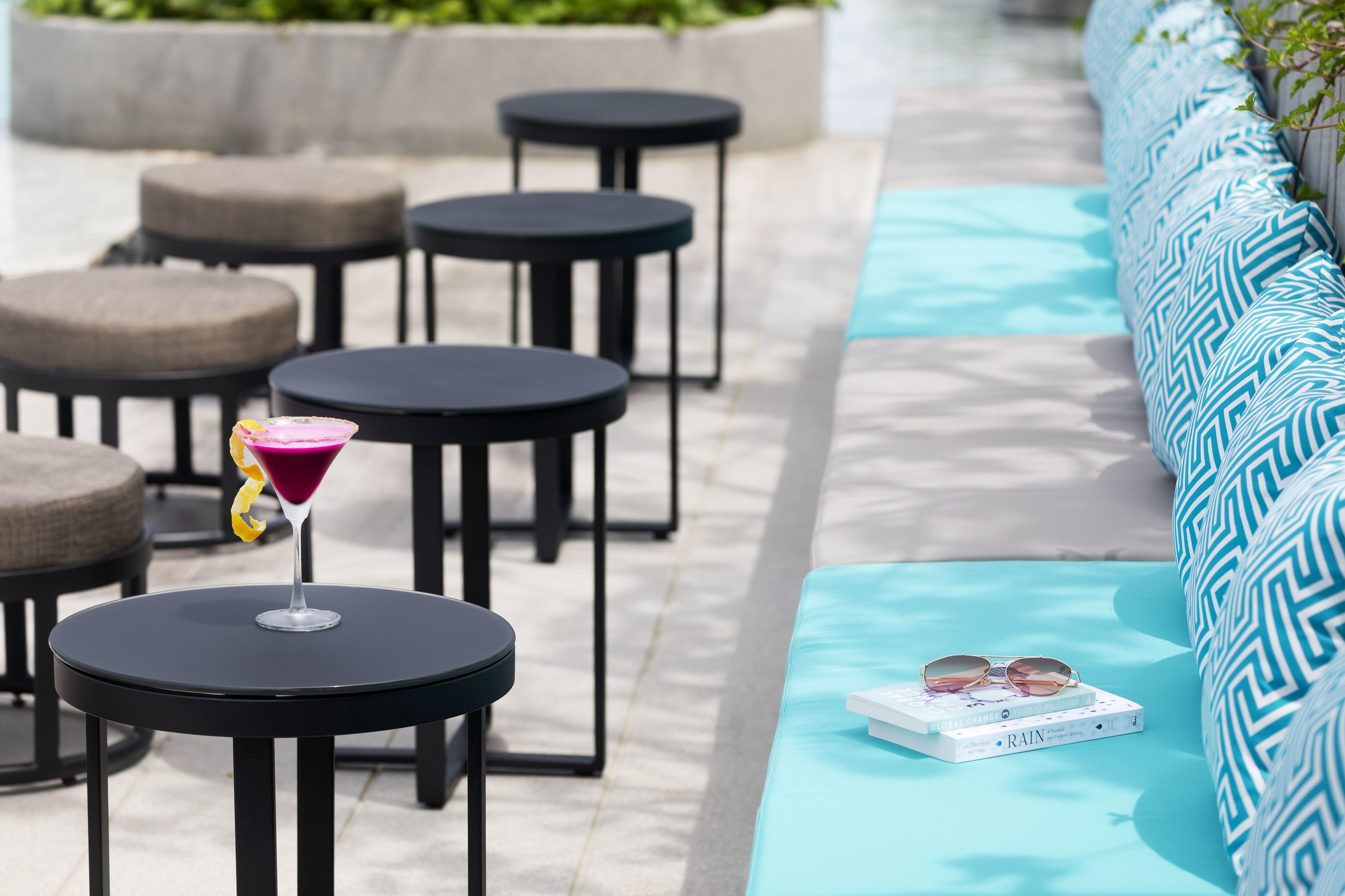 Un espace aménagé au bord de la piscine, avec des tables rondes et un canapé. Une boisson est posée sur l’une des tables, et un livre sur le canapé.