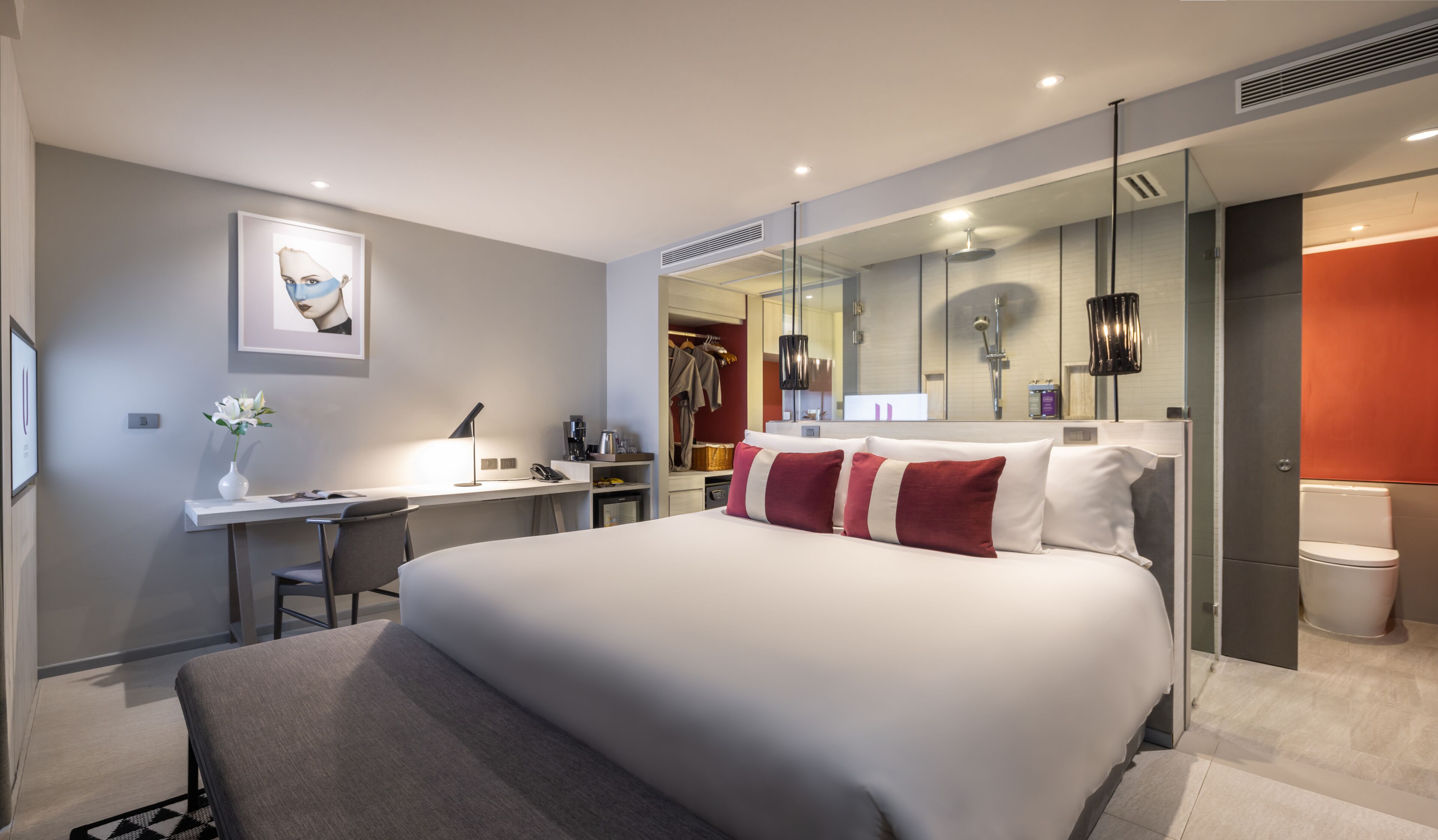A modern hotel room with a large bed, a desk with a chair, a wardrobe, and a bathroom with a toilet and shower.