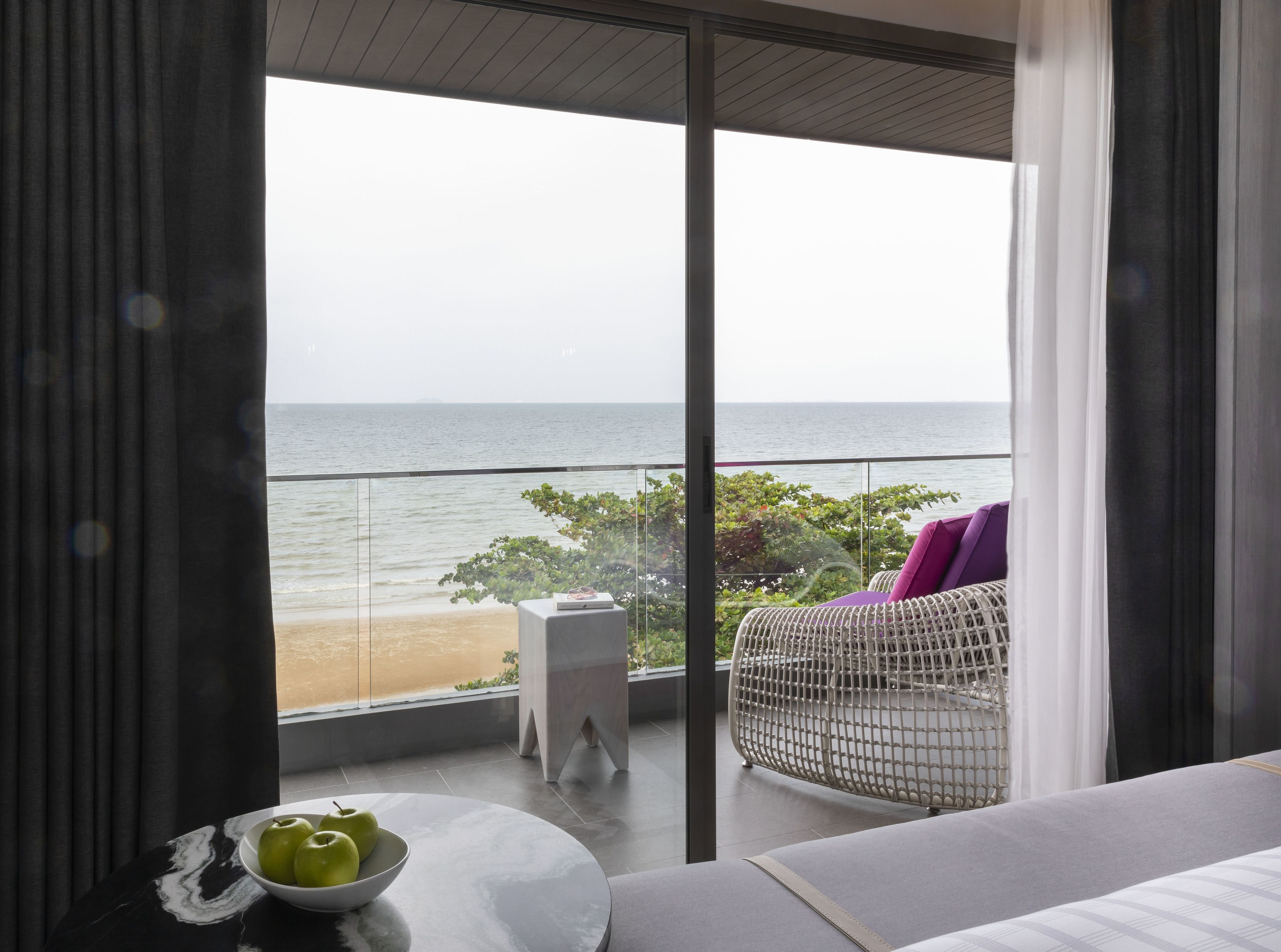Habitación moderna con un amplio ventanal que da a una playa. Hay una silla de mimbre con un cojín morado, una pequeña mesa con un cuenco de manzanas verdes y un sofá blanco.