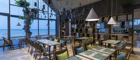Un restaurant moderne avec des tables dressées pour le service, de grandes fenêtres offrant une vue sur la mer et des lampes suspendues décoratives.