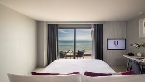Een hotelkamer met een groot bed, een bureau met een stoel, een kleine tafel met stoelen, uitzicht op de oceaan en een lijst aan de muur.
