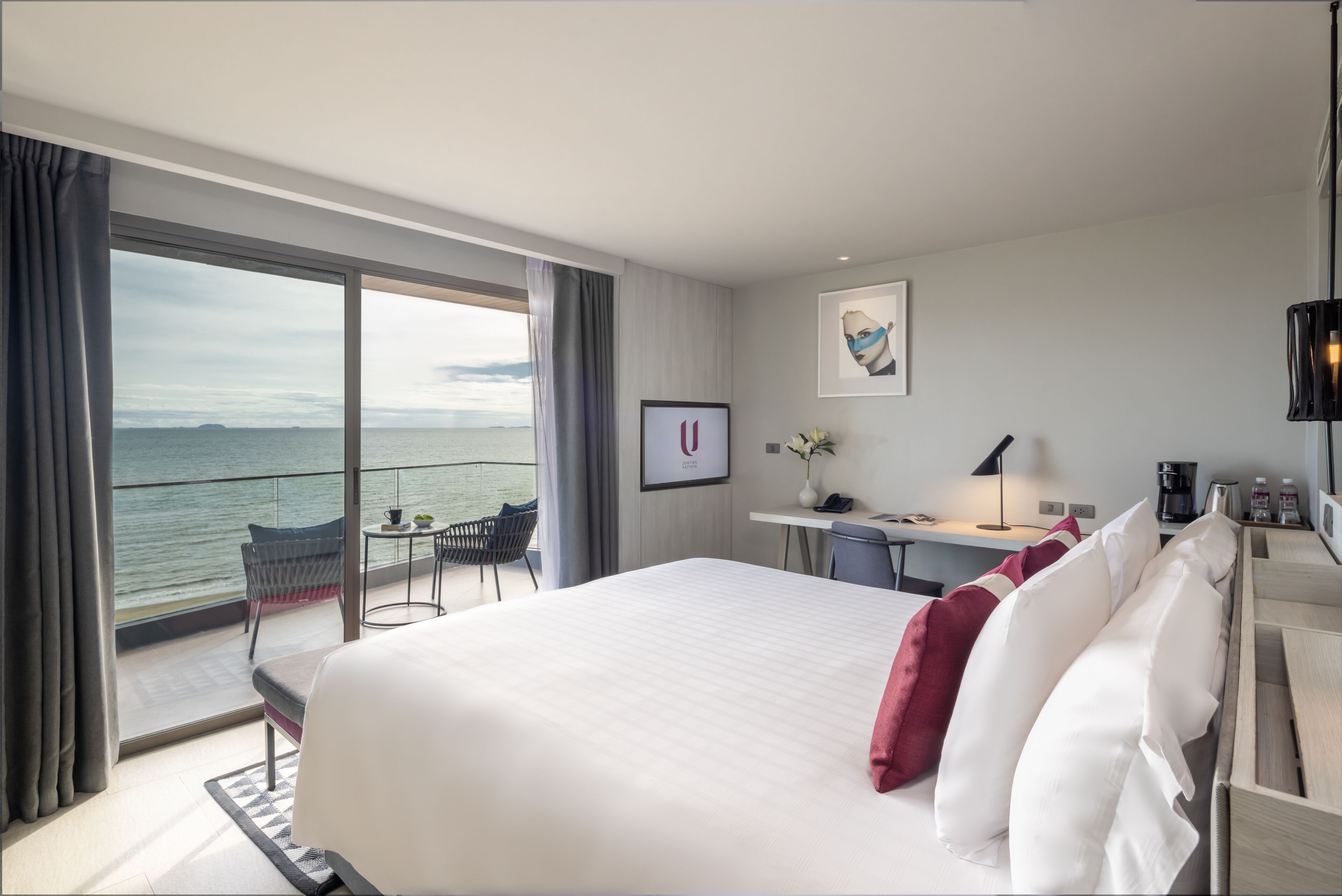A hotel room with a large bed, a desk, a chair, and a balcony with a view of the sea.