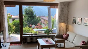 A cozy living room with a wooden table, chairs, and a sofa. A large window offers a view of greenery and buildings. - Haus Dippold (Reit im Winkl)