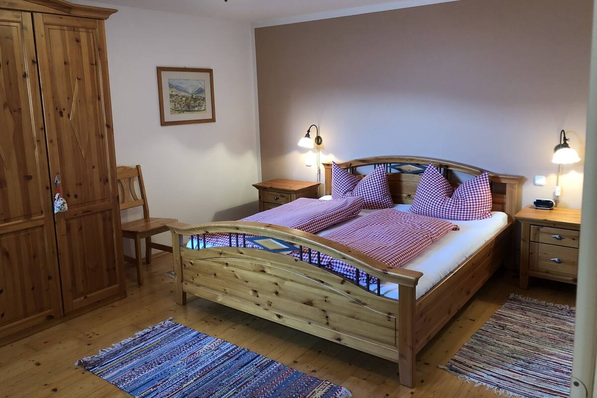 Un lit en bois avec une literie à carreaux, deux lampes de chevet, une table de chevet en bois, une chaise, une armoire en bois et un tableau encadré accroché au mur.