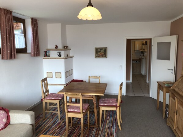 Un coin repas avec une table et des chaises en bois, un tapis à motifs, un meuble blanc et, au fond, une cuisine.
