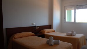 Quarto de hotel com duas camas de solteiro, mesa de cabeceira, luminária e janela com persianas.