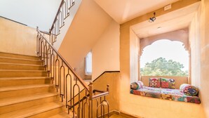 A staircase with a metal railing, a window seat with cushions, and a view of greenery outside. - The Legend Haveli (Jaisalmer)