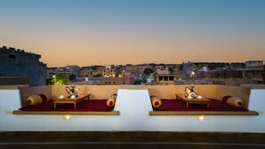 A rooftop terrace with two seating areas, each with a table, cushions, and a hookah. The terrace overlooks a cityscape with buildings in the background. - The Legend Haveli (Jaisalmer)