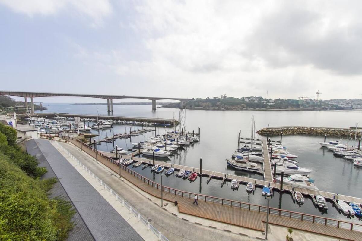 A marina with multiple boats docked, a pedestrian walkway, and a bridge in the background.