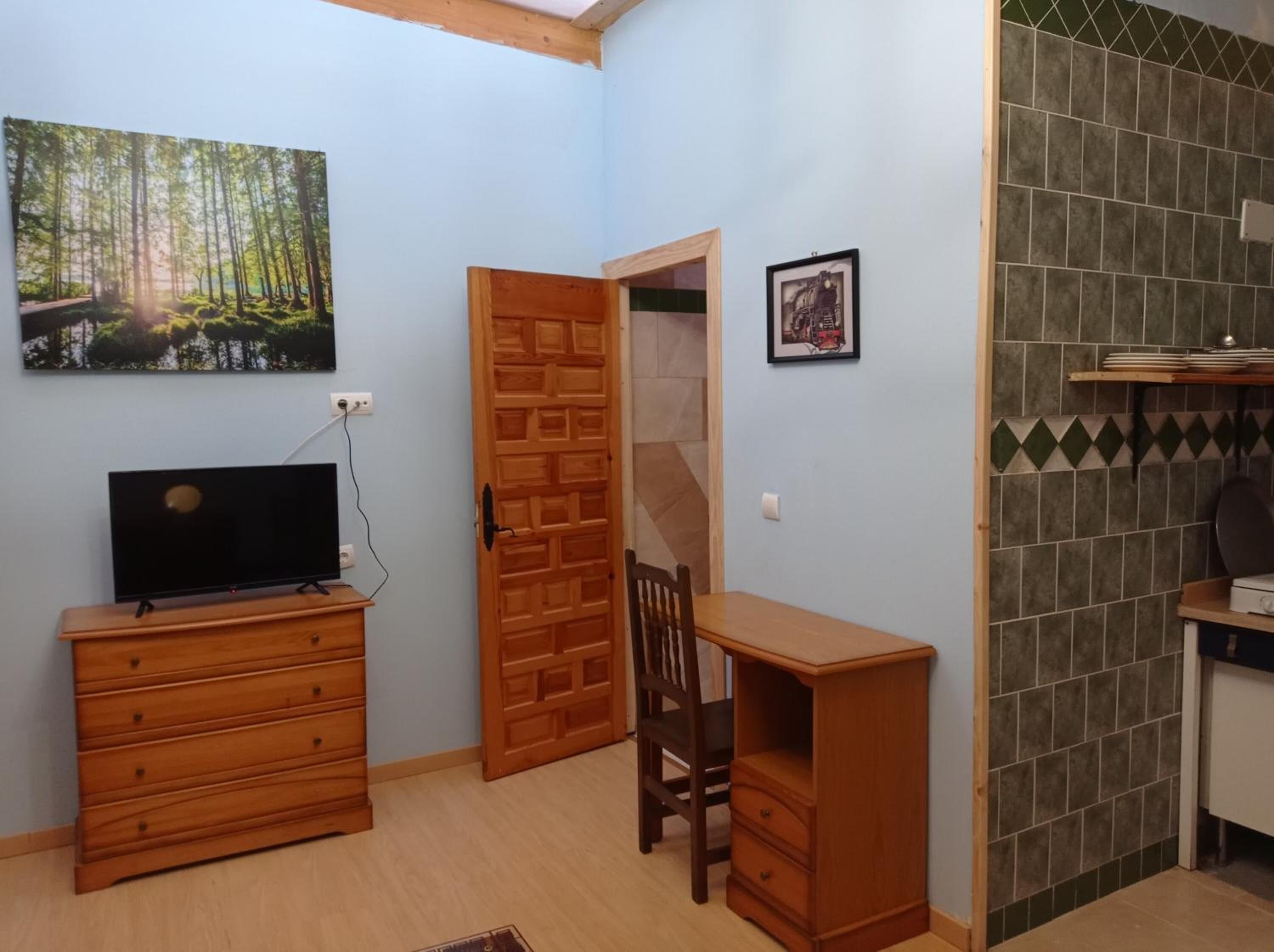 A room with a wooden dresser, a desk, a chair, a television, a painting, and a tiled backsplash.