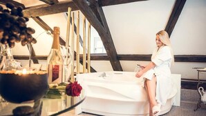 A woman sitting on a white bathtub in a room with a high ceiling and exposed wooden beams. - Schlosshotel Neufahrn (Neufahrn in Niederbayern)