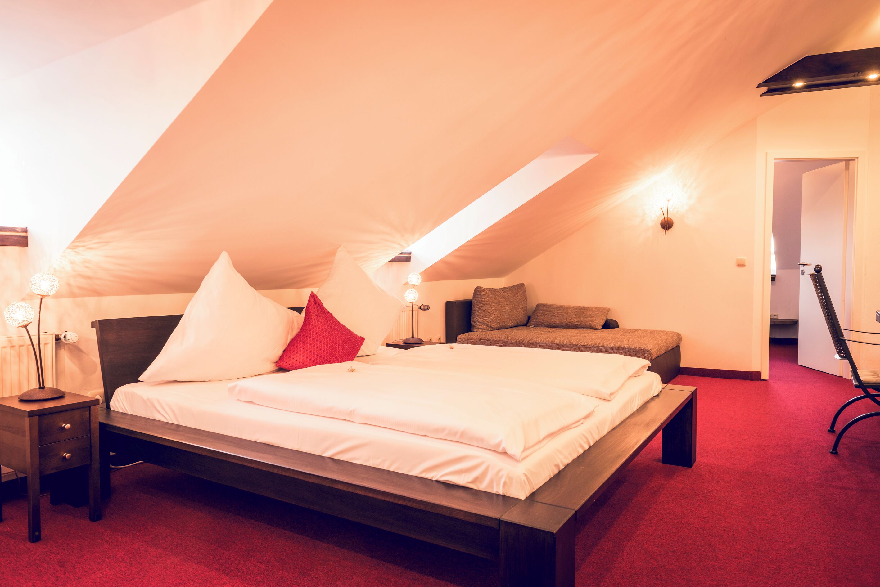 Un dormitorio con techo inclinado, una cama con ropa de cama blanca, una mesita de noche con una lámpara y una alfombra roja.