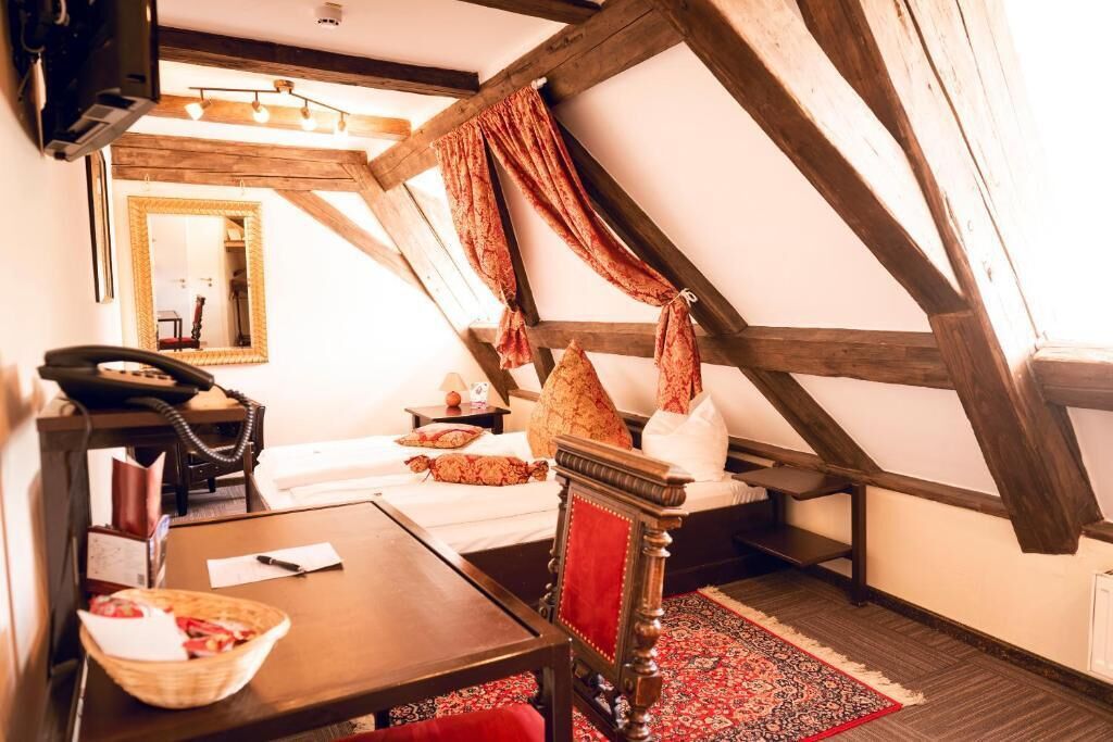 A room with a sloped ceiling, a bed with pillows, a bedside table with a lamp, a chair, and a red rug.