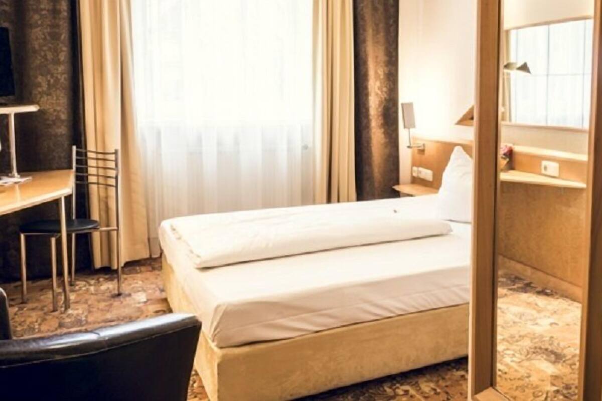 A hotel room with a bed, a desk, a chair, a mirror, and a bathroom with a toilet and sink.