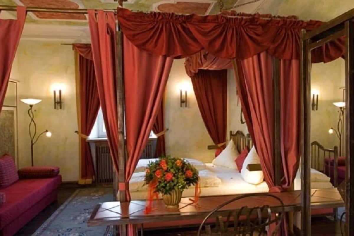 A bedroom with a four-poster bed, red curtains, a bedside table with a vase of flowers, and a red sofa.