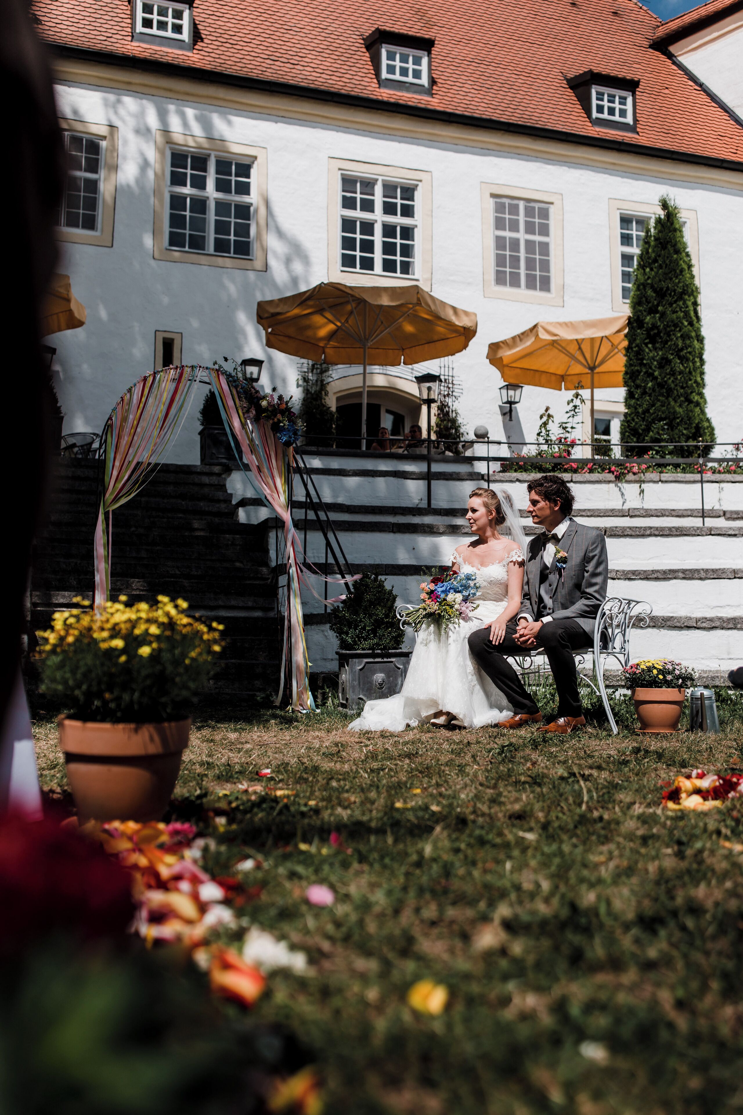 A wedding ceremony with a couple seated on a bench in front of a building with a red-tiled roof and white walls.