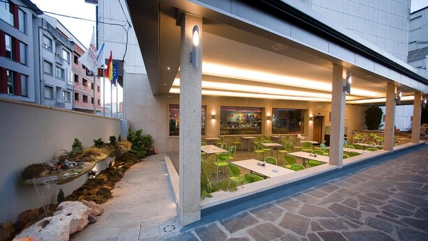 An outdoor dining area with stone flooring, green chairs, and a view of the restaurant inside. - Hostal Terra Chá (Villalba)