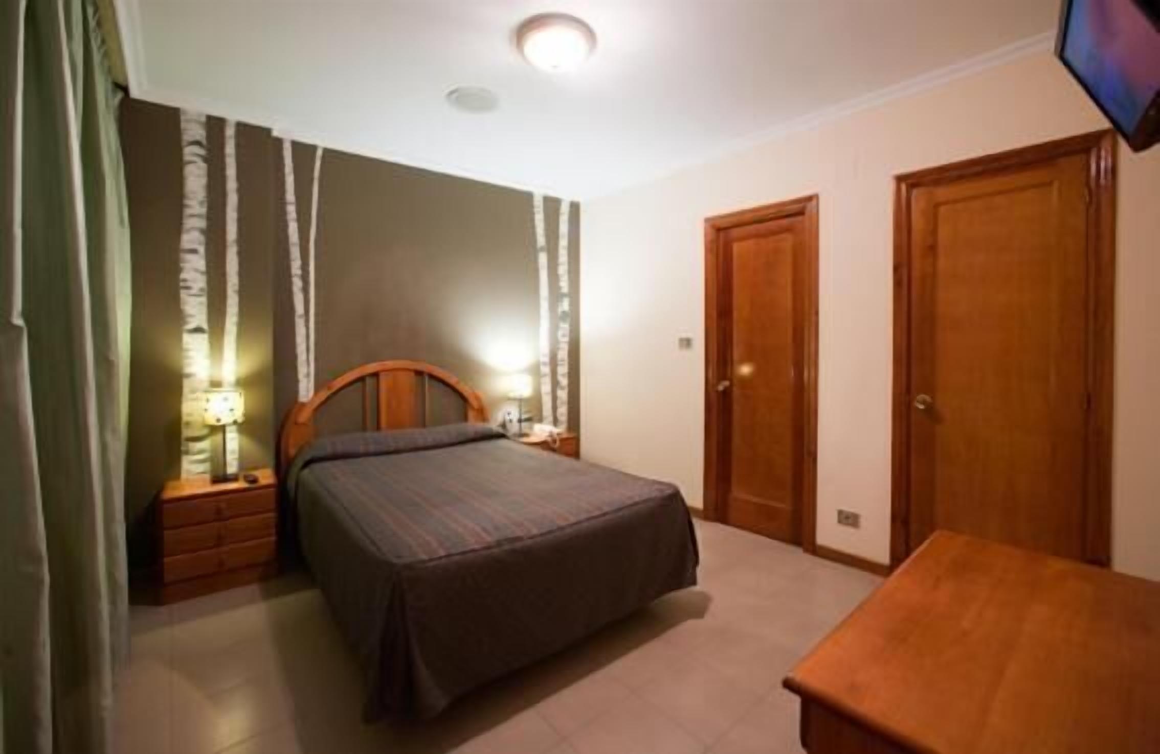 Un dormitorio con cama, mesitas de noche, un banco de madera y dos puertas.