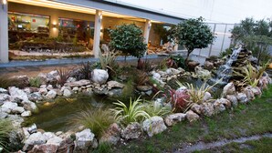 A landscaped garden with a pond, rocks, and plants in front of a building with large windows and a covered walkway. - Hostal Terra Chá (Villalba)