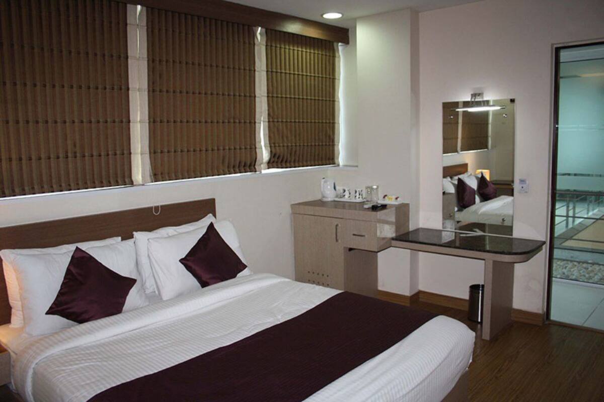 A hotel room with a bed, bedside table, mirror, and window with blinds.