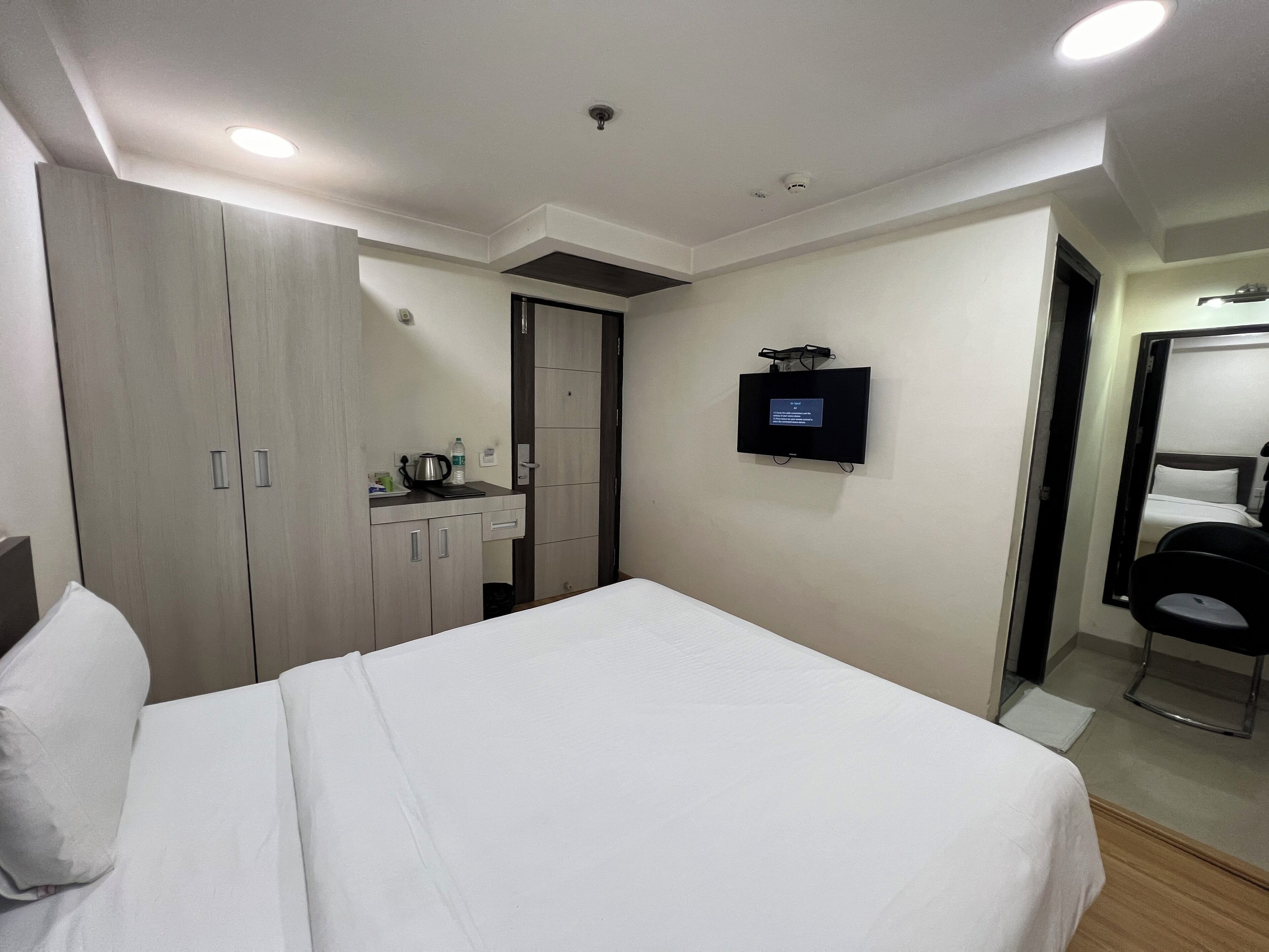 A hotel room with a bed, a TV mounted on the wall, a desk with a kettle, and a wardrobe.