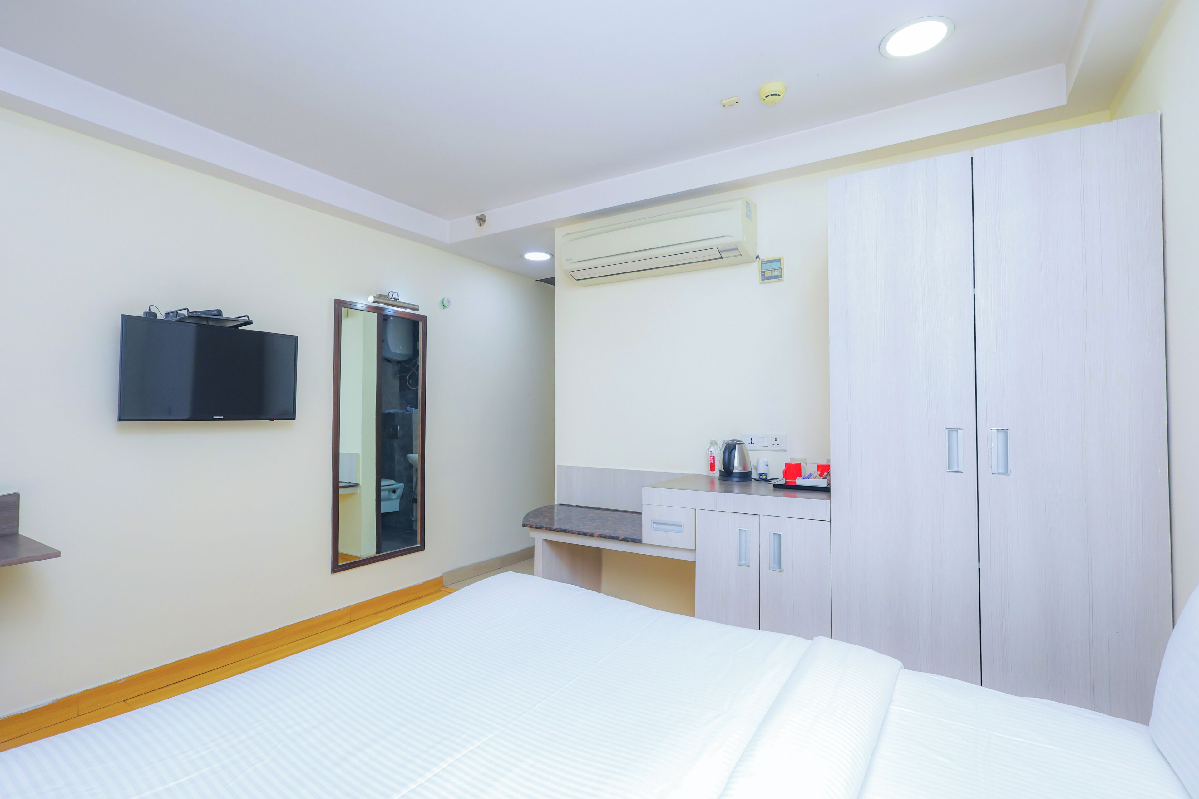 A hotel room with a bed, a mirror, a wardrobe, a small desk with a kettle, and an air conditioning unit.
