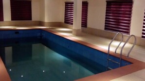 A rectangular indoor swimming pool with a ladder on one side, surrounded by beige walls and windows with dark blinds. - Reeve Court Apartment (Lagos)