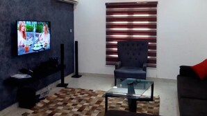 A modern living room with a flat-screen TV, a dark armchair, a glass coffee table, and a patterned rug. - Reeve Court Apartment (Lagos)