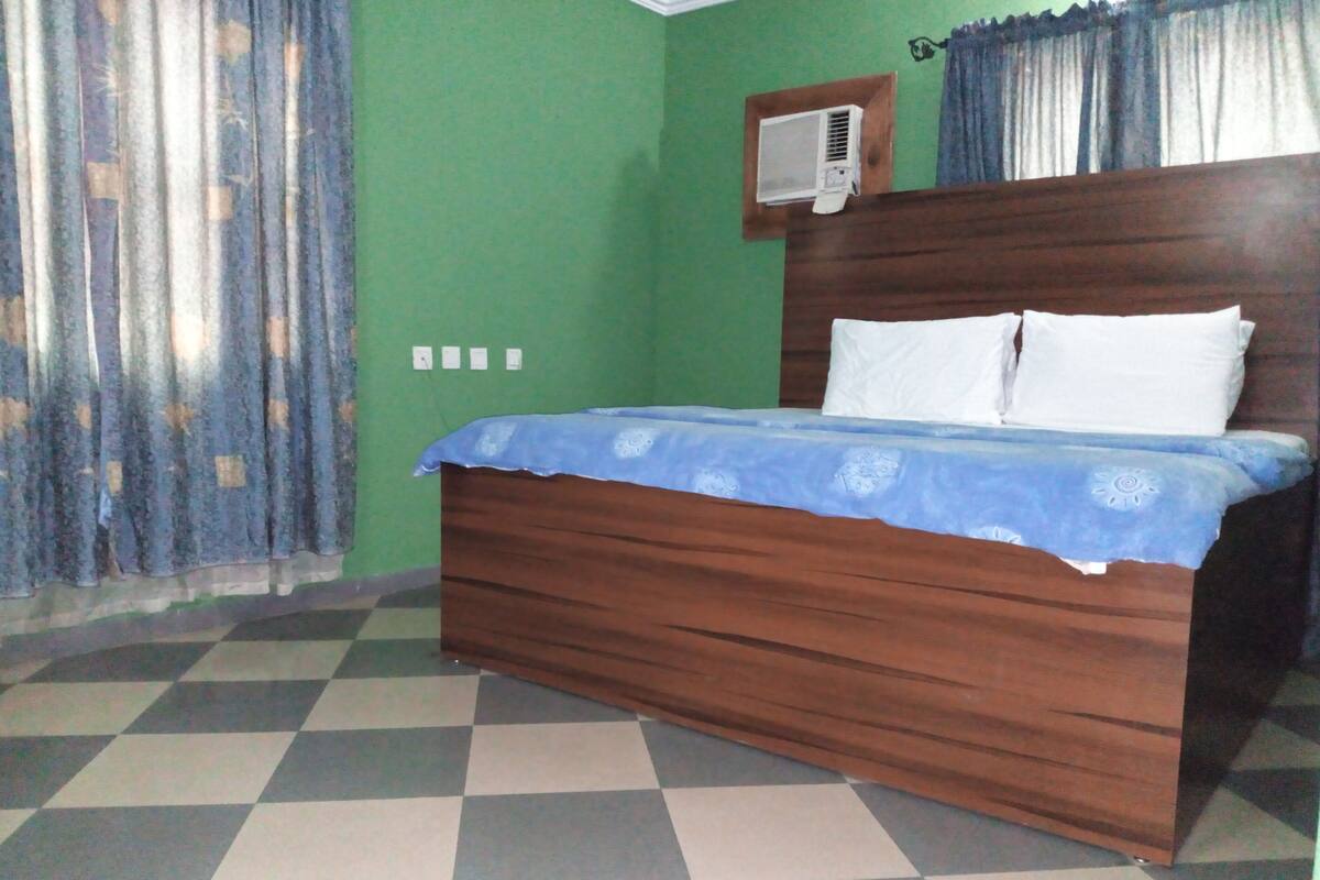 Ein Zimmer mit kariertem Boden, einem hölzernen Bett mit blauer Bettdecke, grünen Wänden und einem Fenster mit Vorhängen.