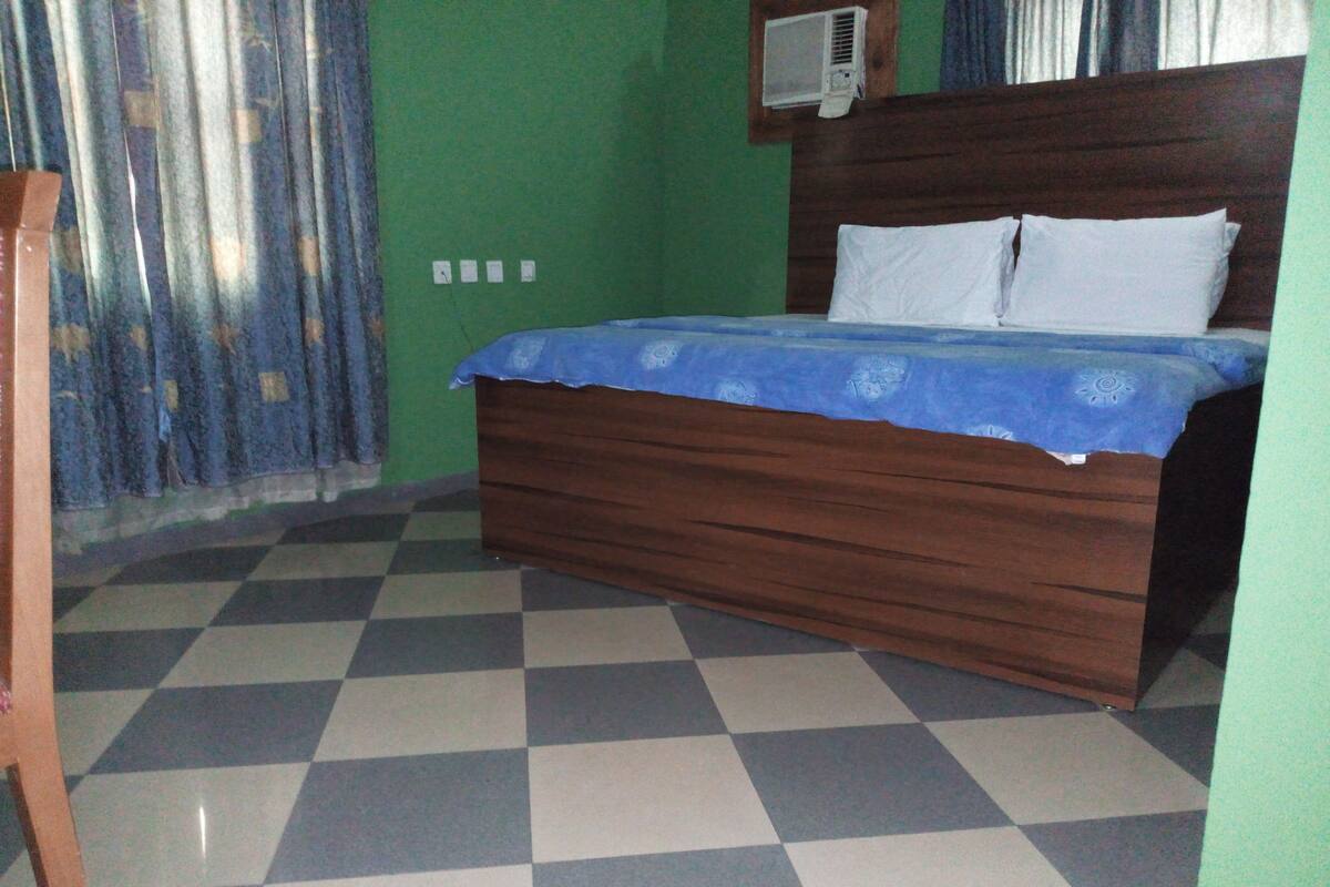 Ein Zimmer mit Schachbrettmuster-Boden, einem hölzernen Bett mit blauer Bettdecke, weißen Kissen und einer Wand-Klimaanlage.