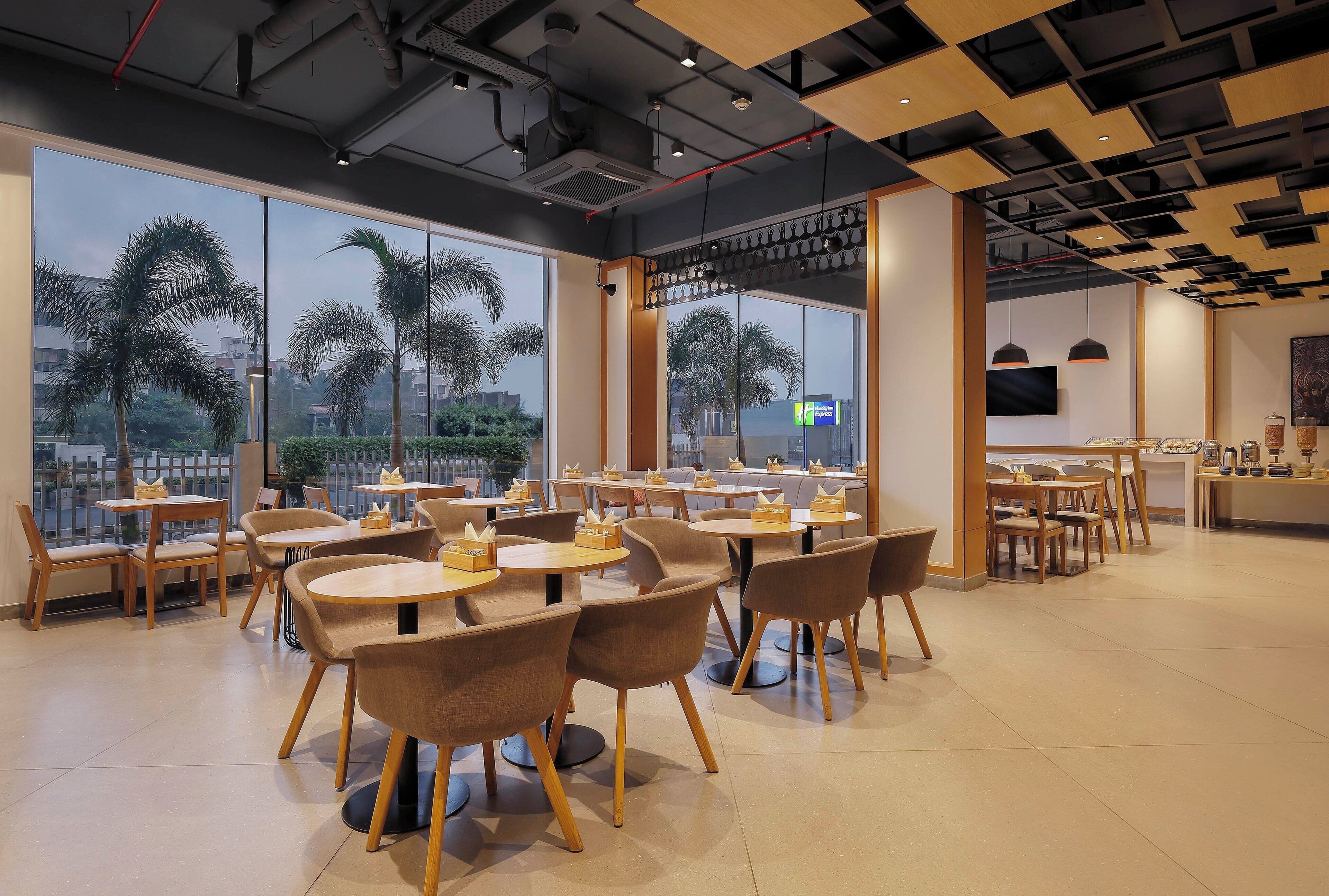 A spacious cafe with large windows, wooden tables, and chairs, and a view of palm trees outside.